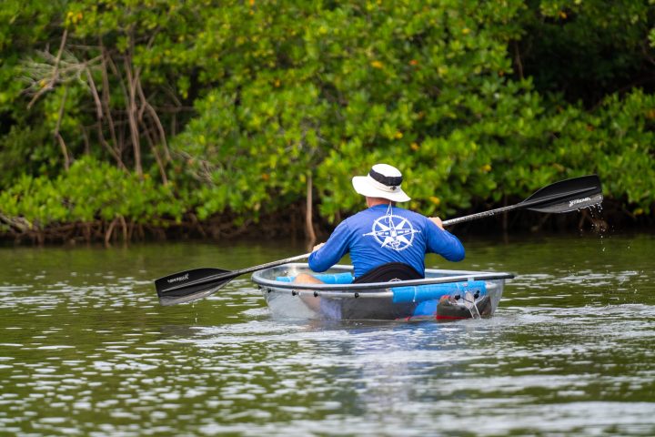 Person paddling in a round, transparent kayak on a calm river.