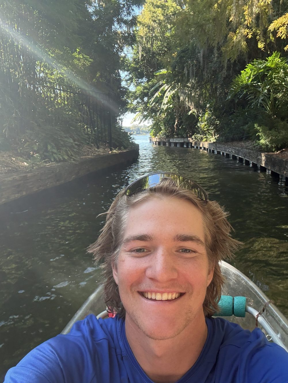 Person smiling in a boat on a lush green canal under sunlight.
