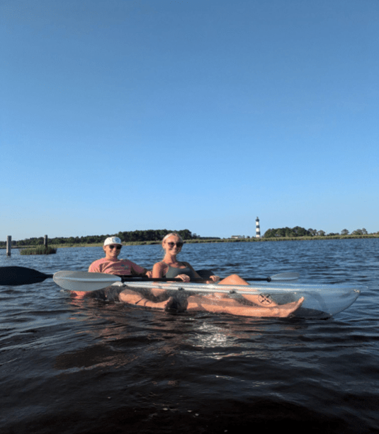 Two people kayaking in a clear kayak on a lake with distant lighthouse.