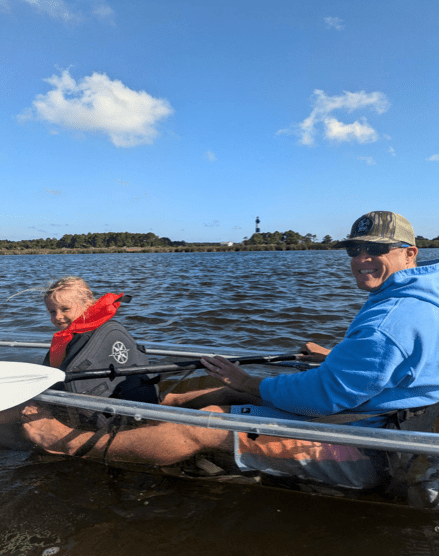 Two people kayaking on a lake with a distant lighthouse and clear sky.