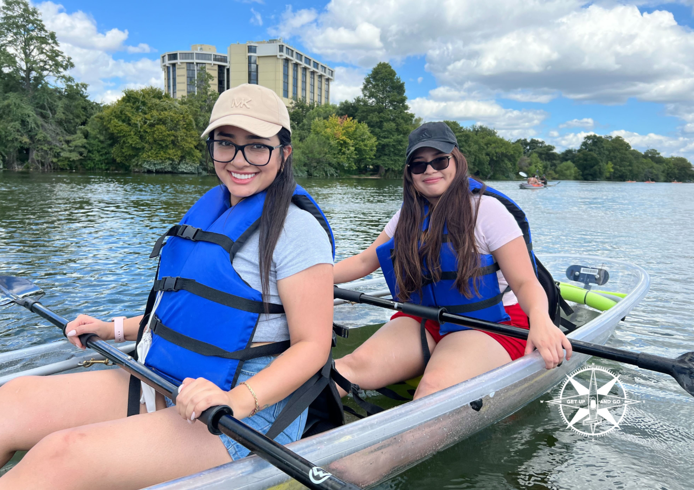 Two women kayaking on a lake, wearing blue life jackets under a partly cloudy sky.