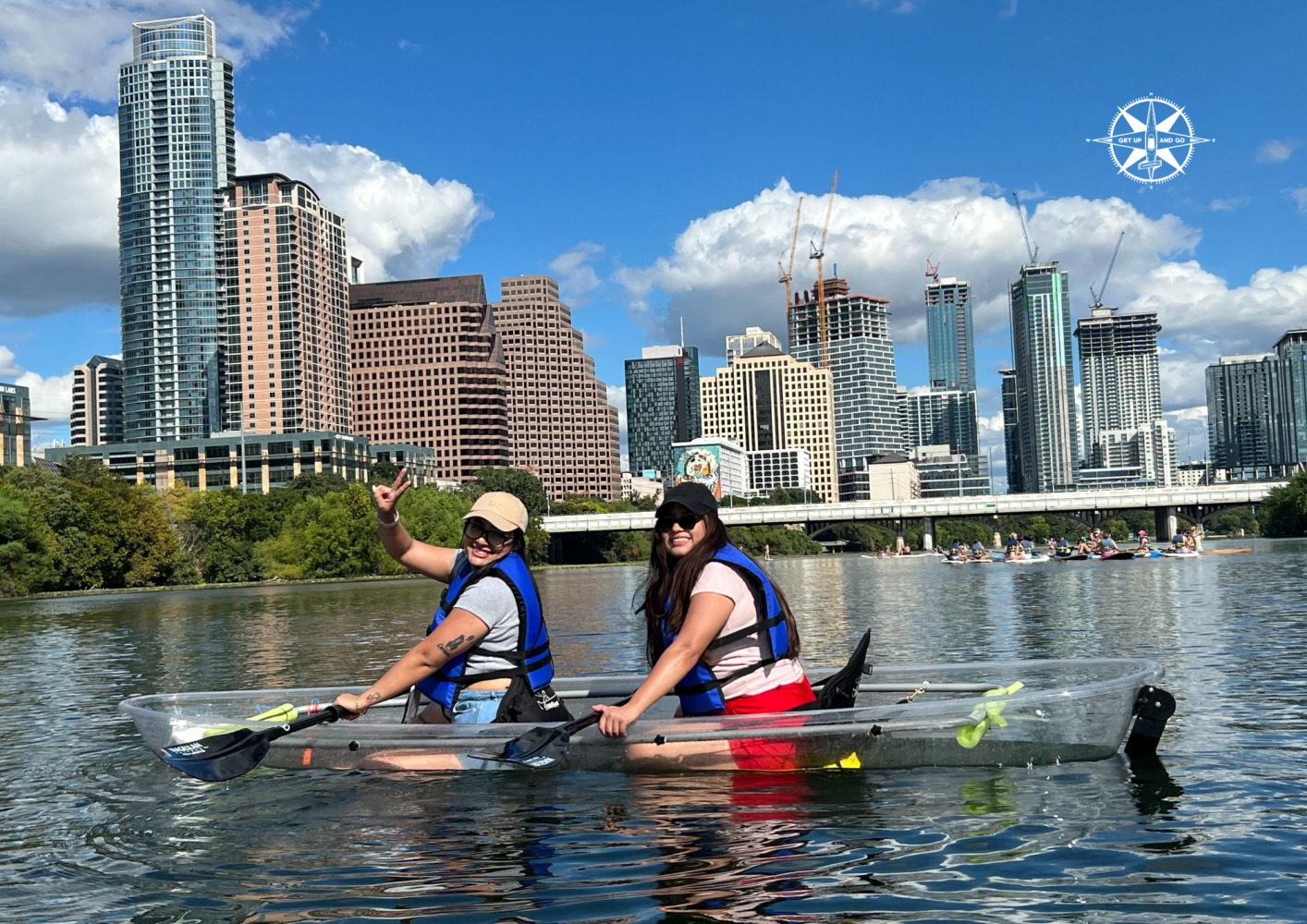 Two people kayak on a river with a city skyline and blue sky background.