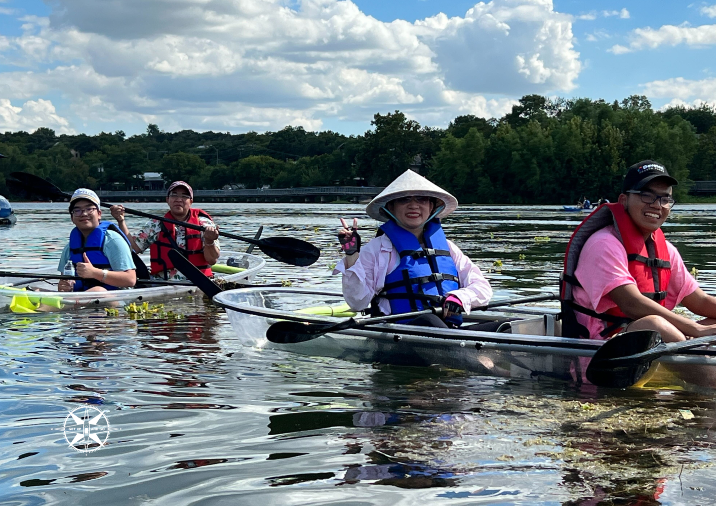 Four people in clear kayaks on a lake, wearing life vests and smiling under a partly cloudy sky.