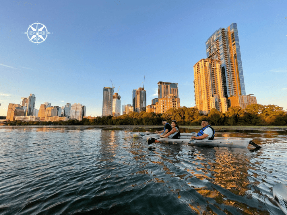 Three people kayaking on a river with city skyline in the background during sunset.