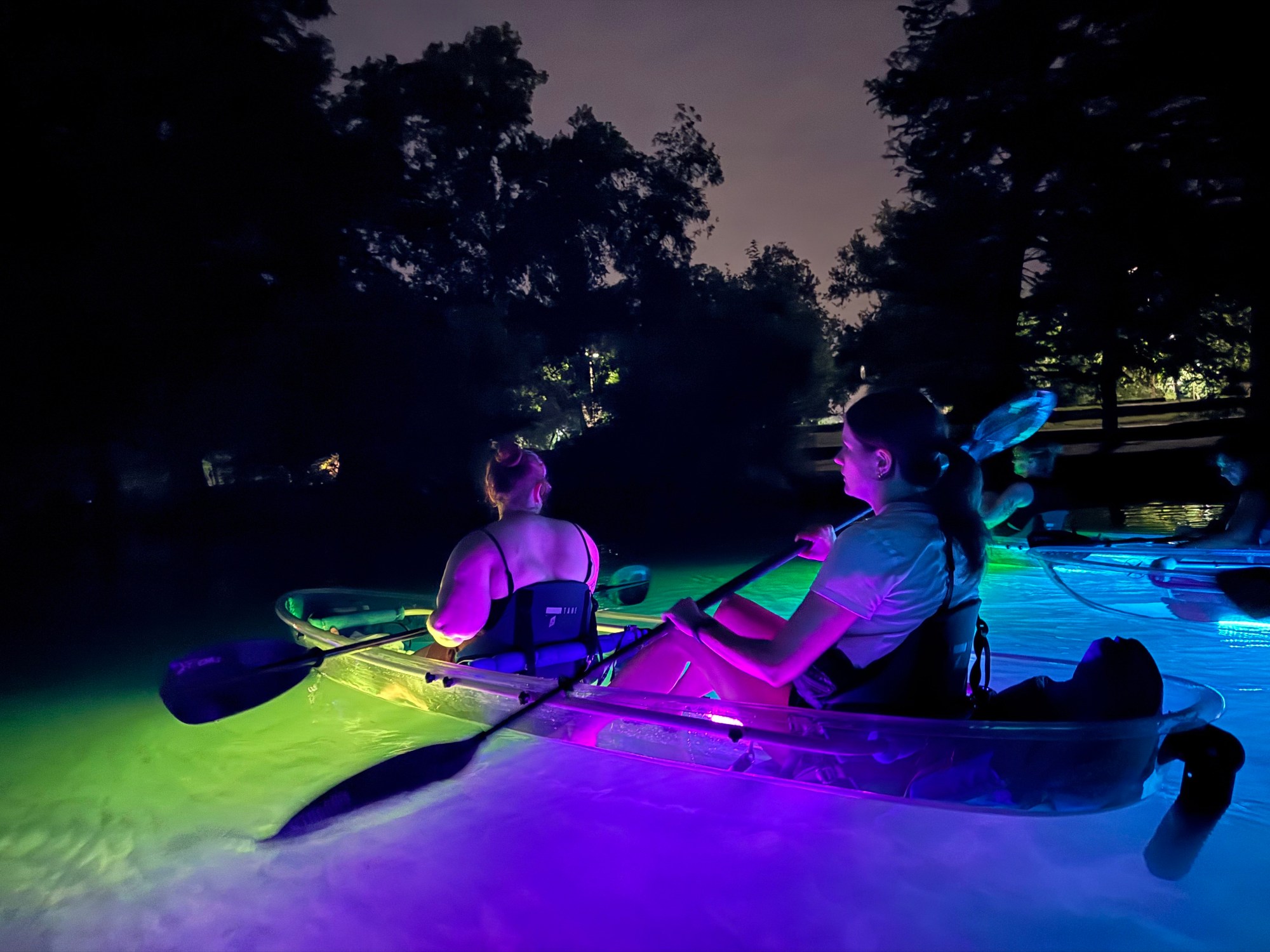 IMG_3808 Two people kayaking at night, illuminated by colorful lights on the water.