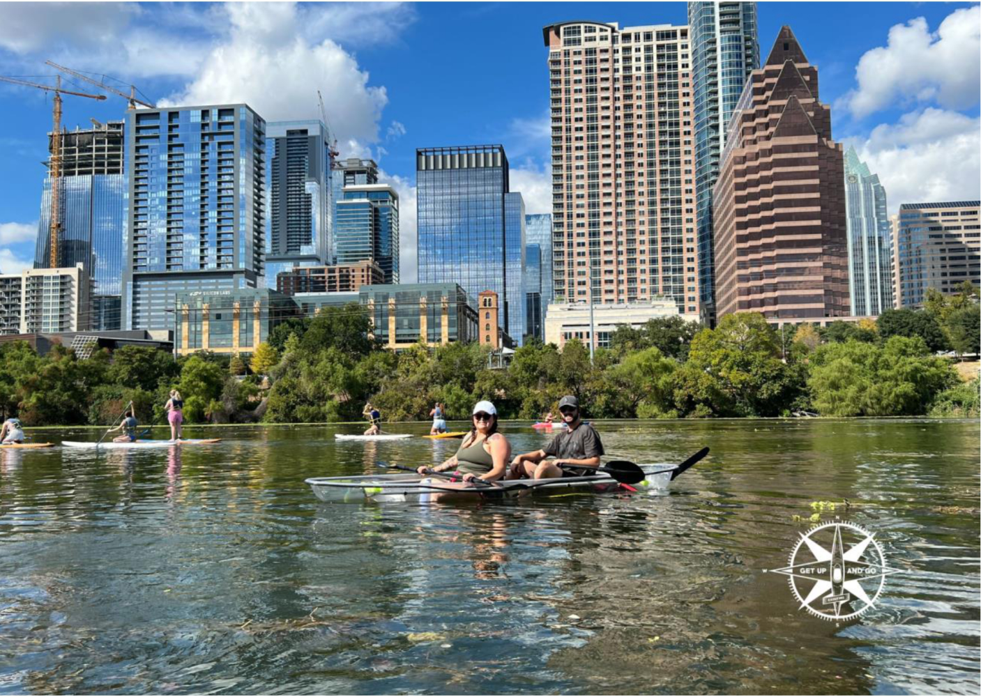 People kayaking on a river with a city skyline in the background during a sunny day.