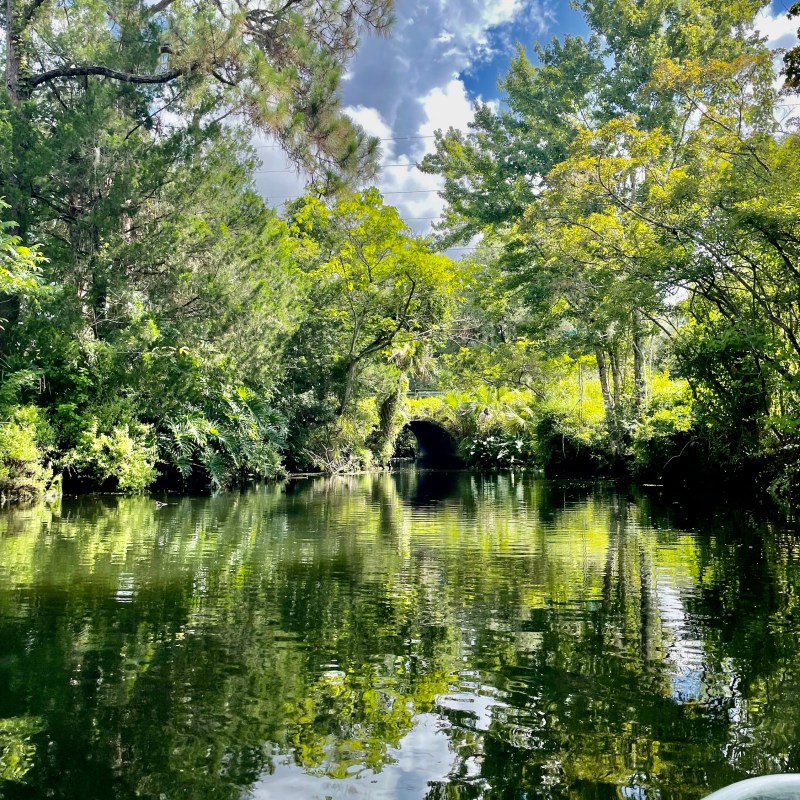 Scenic waterway with lush trees and stone bridge under a partly cloudy sky.