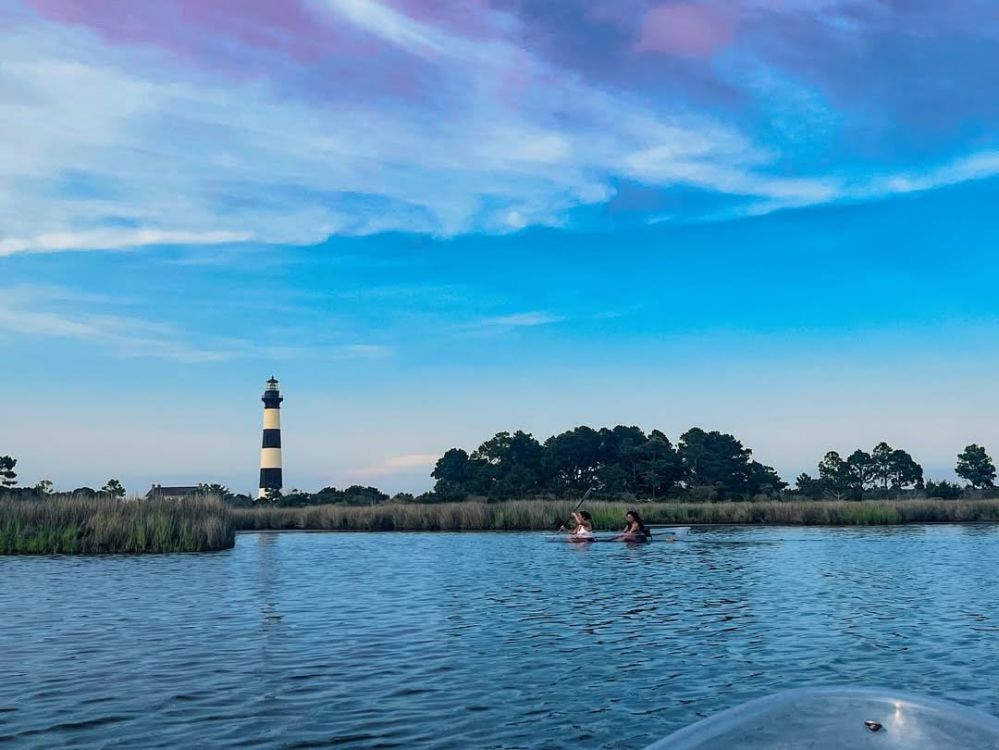 Two kayakers on water near a lighthouse and trees under a blue sky.