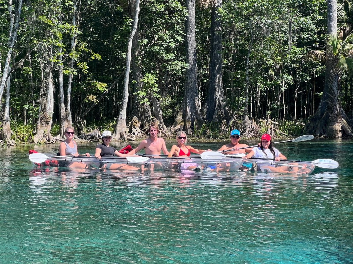 Group of people kayaking in clear kayaks on a calm river surrounded by trees.