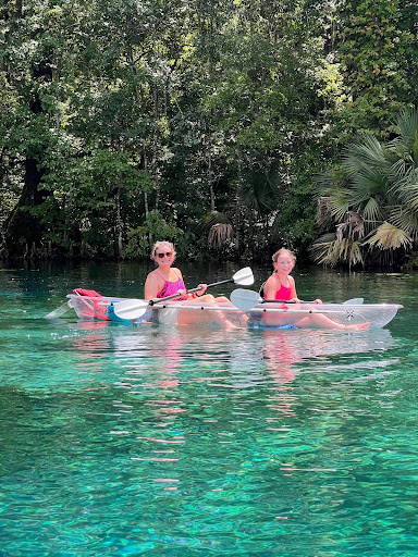 Two people kayaking on clear water with trees in the background.