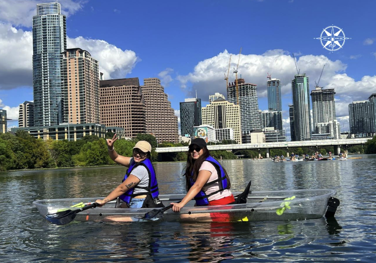 Two people in a clear kayak on a river with a city skyline in the background.
