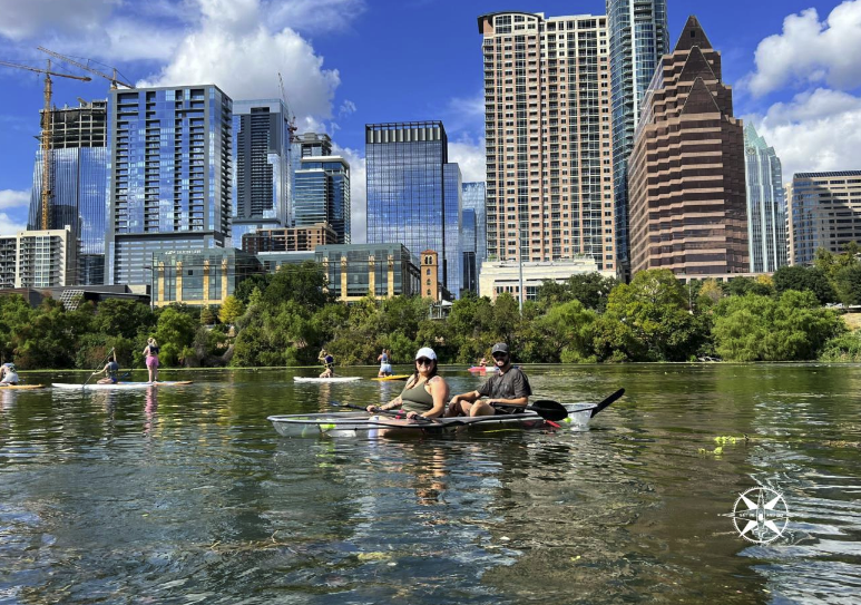 People kayaking on a river with city skyscrapers in the background on a sunny day.