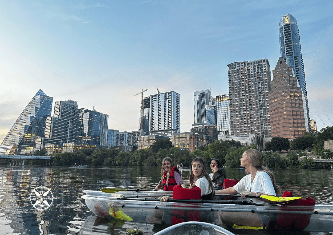 People in kayaks on river with city skyscrapers in background during sunset.