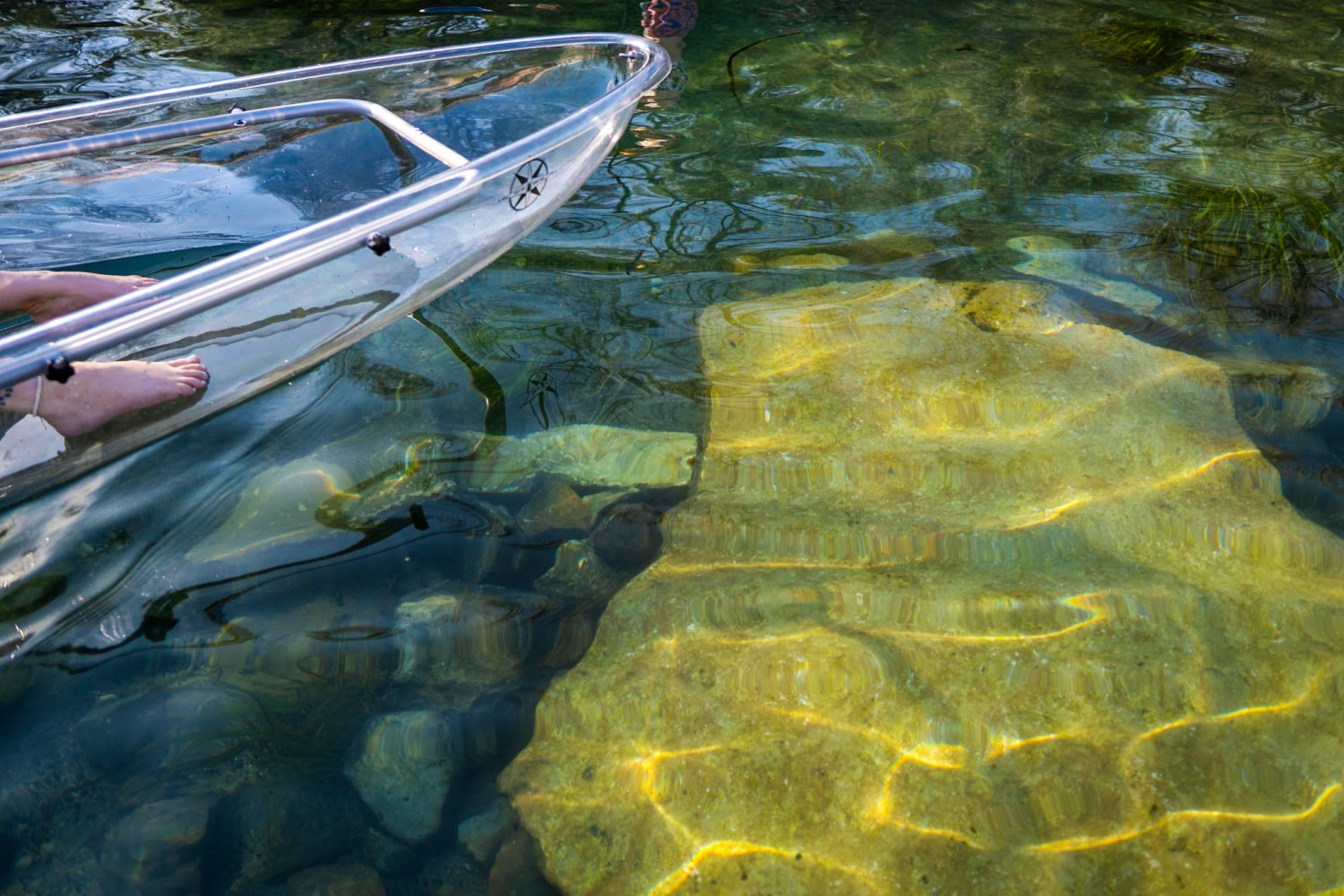 Feet visible in a transparent kayak floating on clear water with rocks below.
