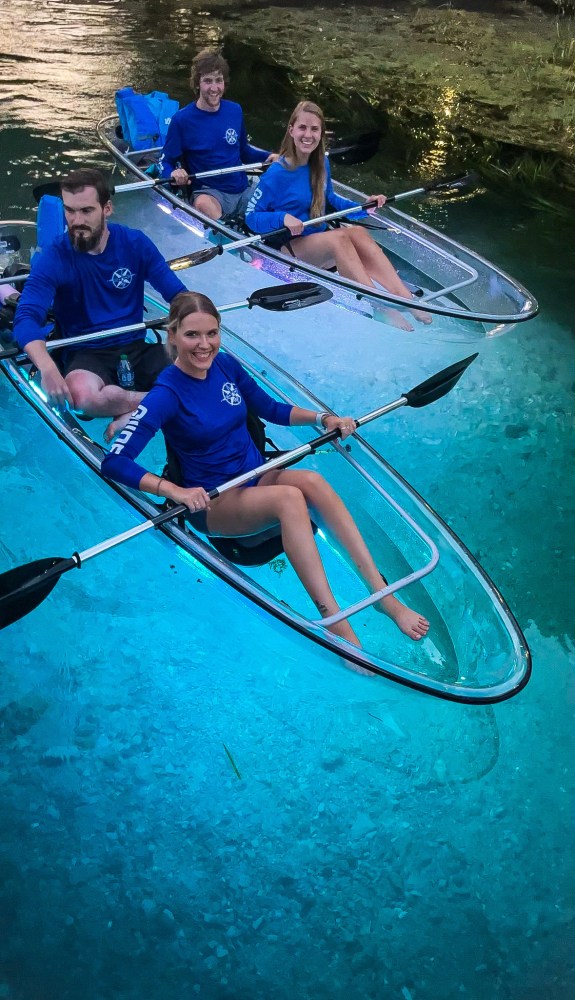 Two clear kayaks on a calm, blue river with four people in blue shirts paddling.