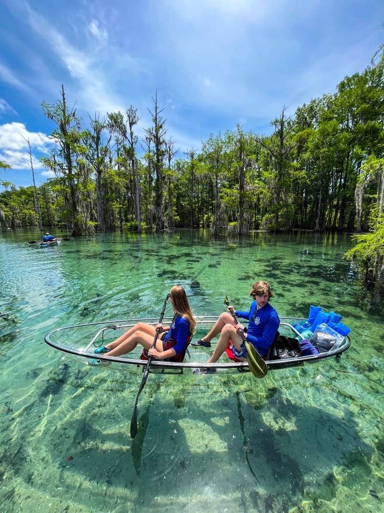 Two people in a transparent kayak on clear water surrounded by trees under a blue sky.