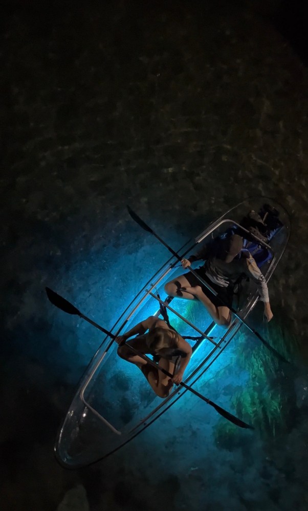 Two people in a clear kayak with blue light paddling at night.