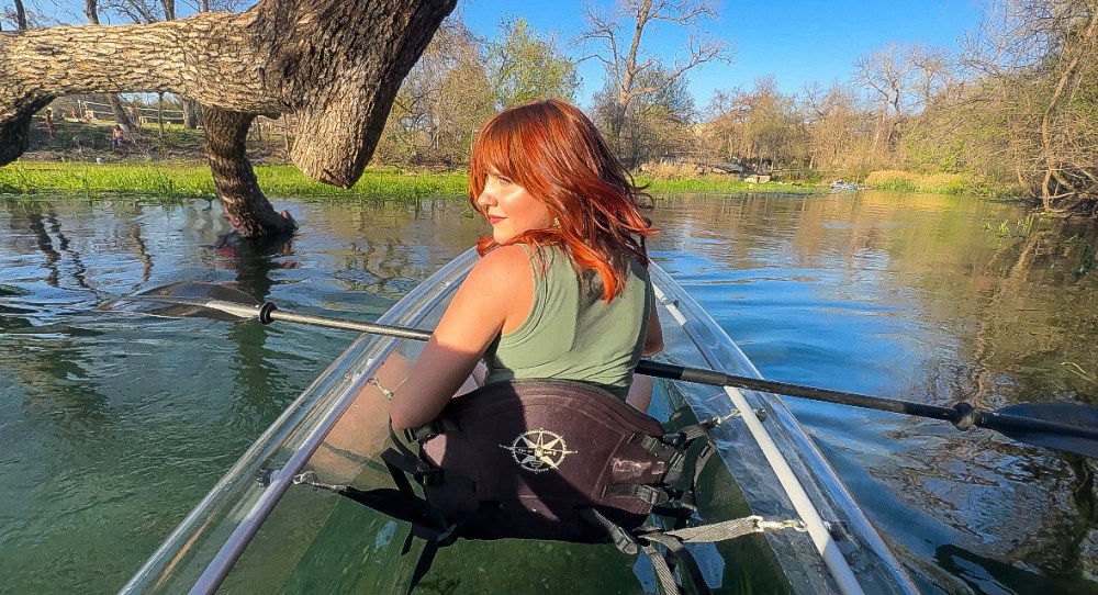 Person with red hair kayaking on a clear waterway lined with trees on a sunny day.