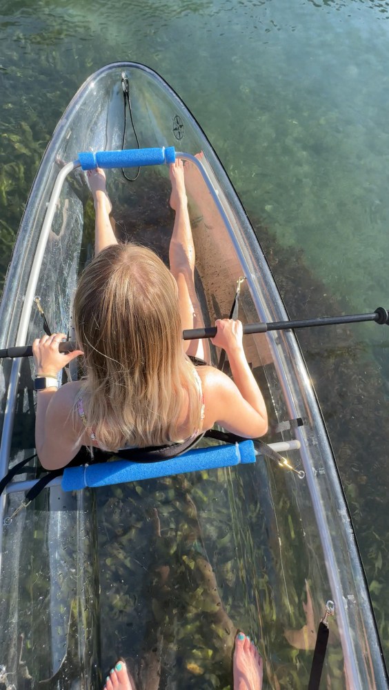 Person paddling a transparent kayak in clear water with submerged plants visible.