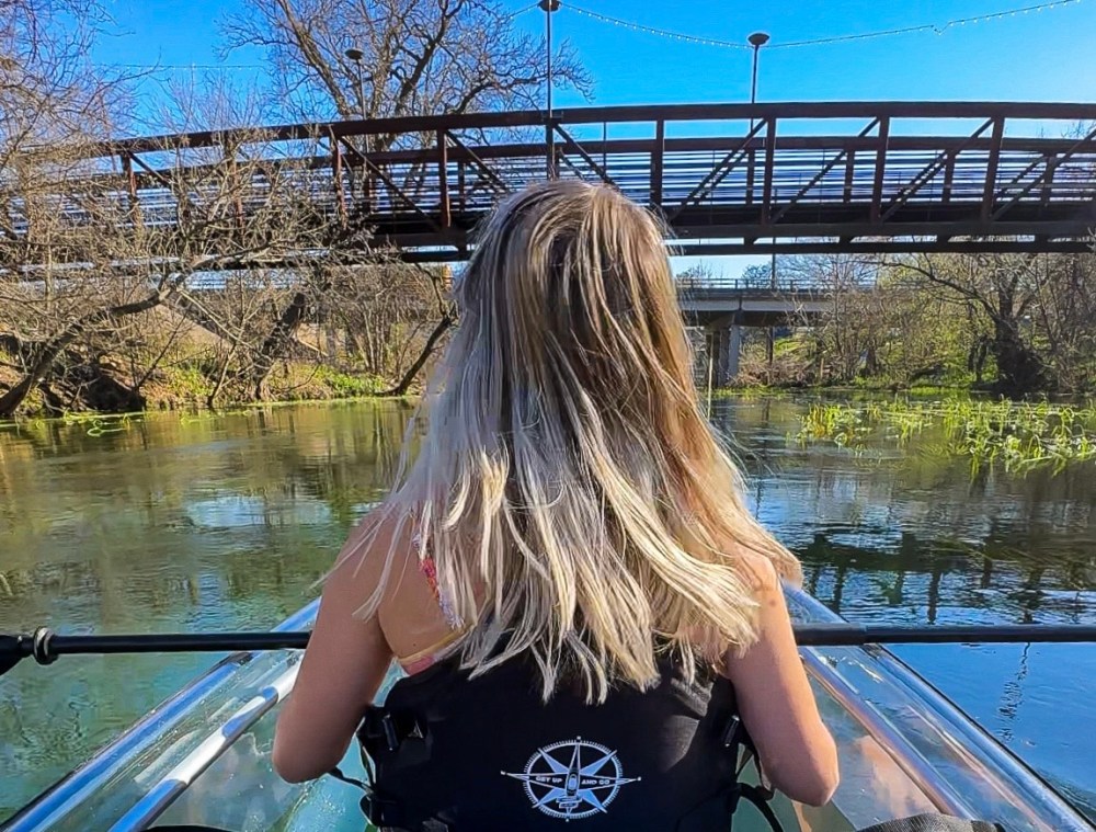 Woman kayaking under a bridge on a sunny day with green surroundings.