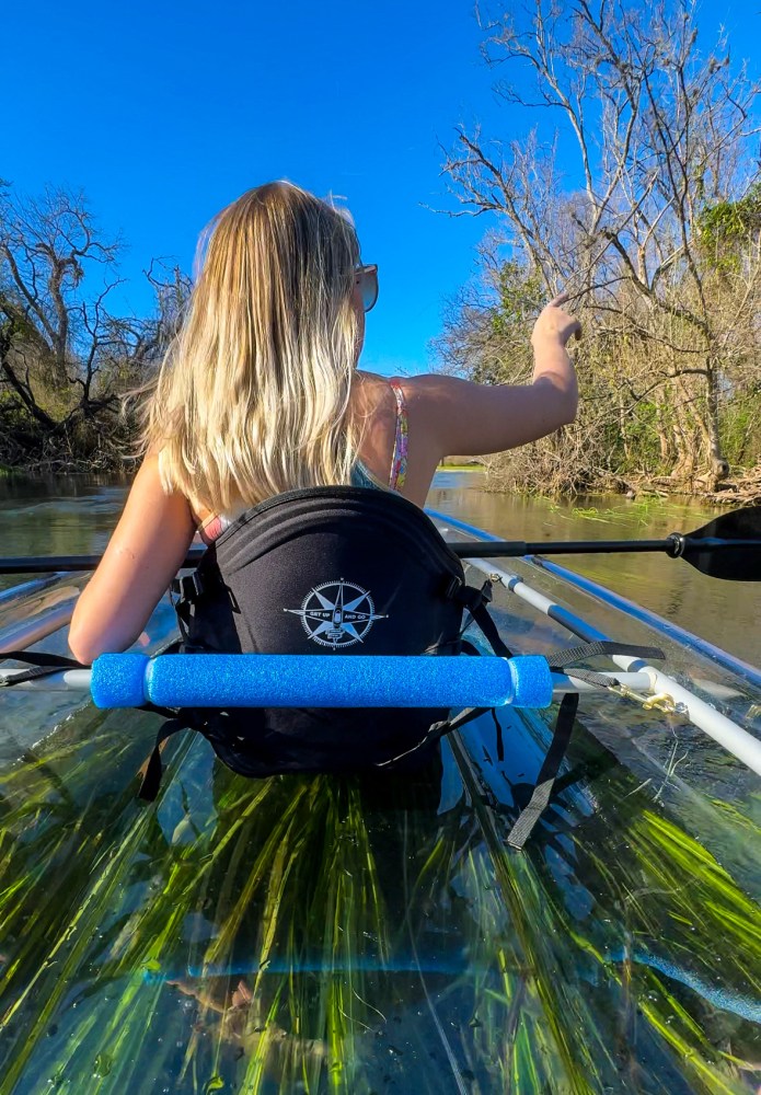 Person with long hair kayaking on clear water under blue sky, pointing at trees.
