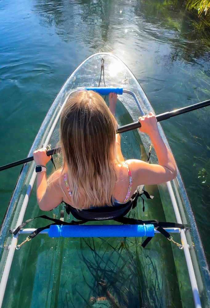 Woman paddling a clear kayak on blue water, view from behind.