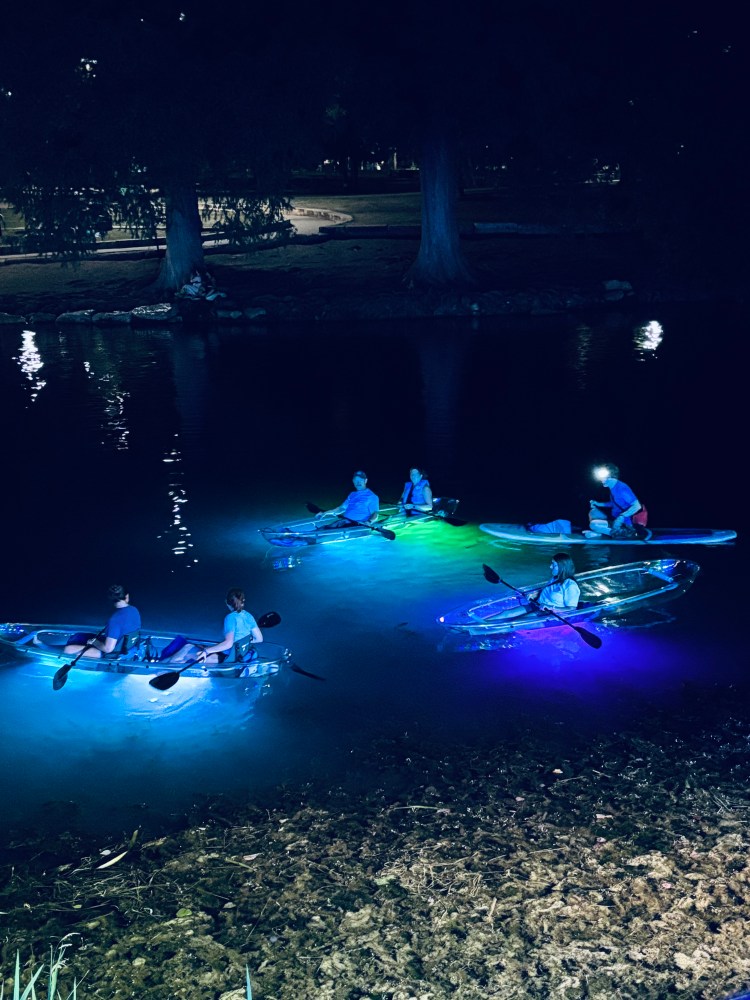 People kayaking at night in transparent kayaks with colorful glowing lights on a calm river.