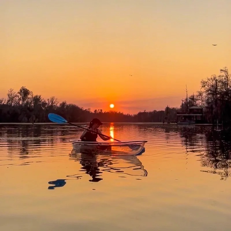 Sunset Kayak Person kayaking on a calm lake at sunset with an orange sky and silhouetted trees.