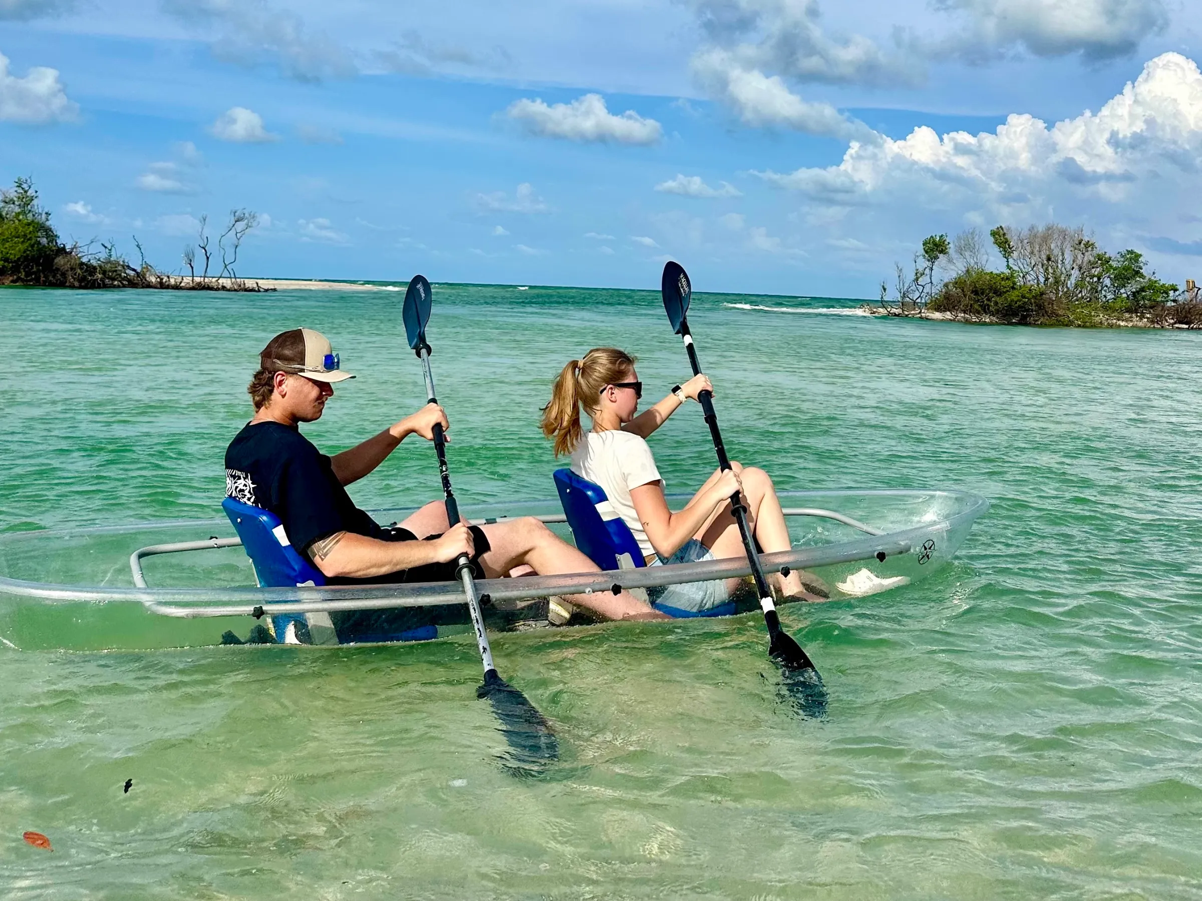 Two people kayaking in a transparent kayak on clear water with distant trees and clouds.