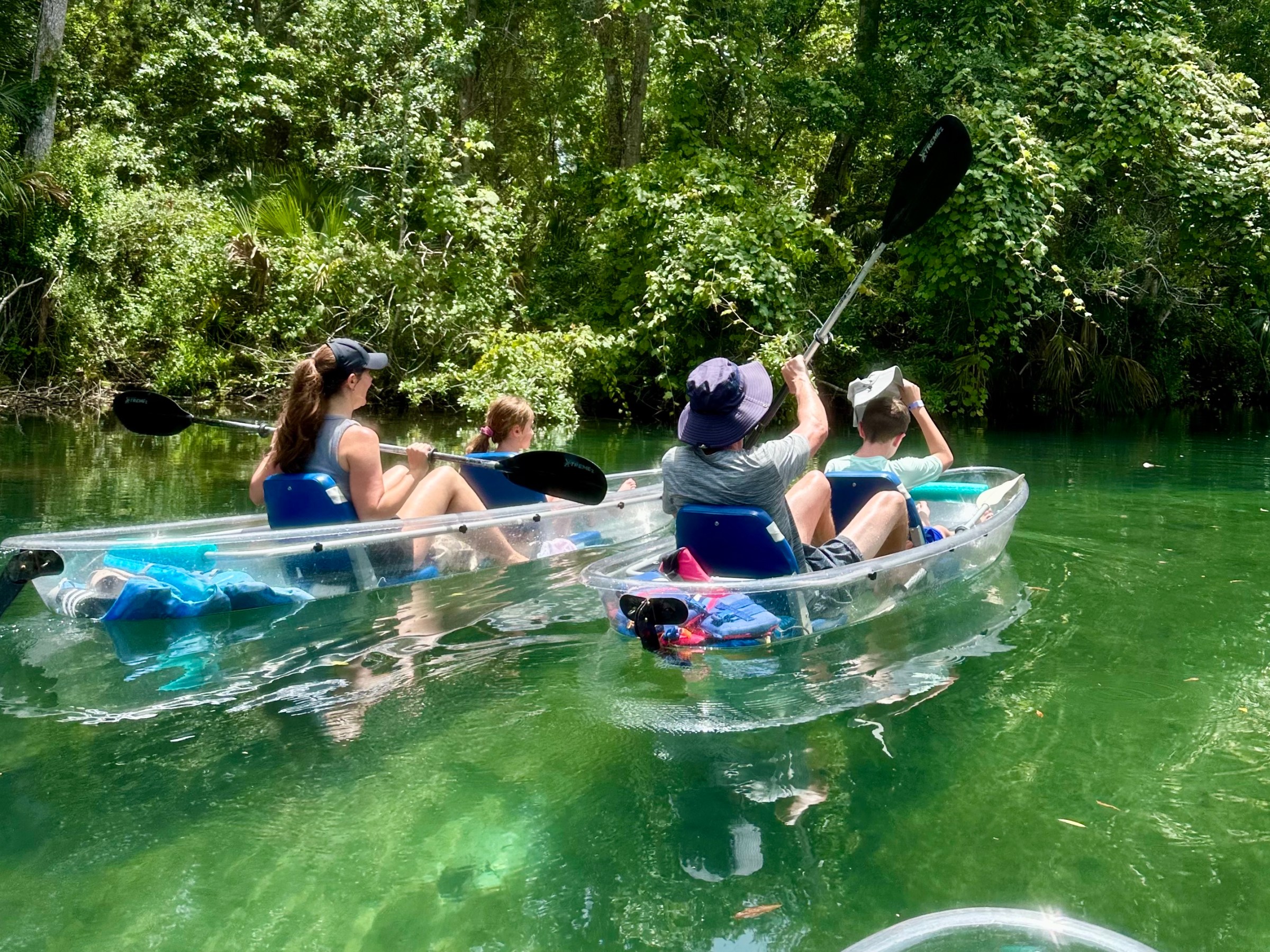 Two clear kayaks with four people paddling on a green river surrounded by lush trees.
