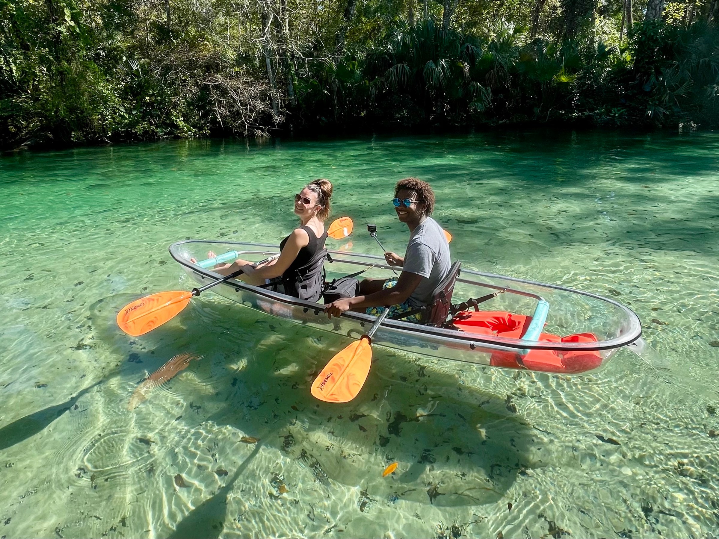 Two people in a transparent kayak on clear, green water with lush greenery in the background.