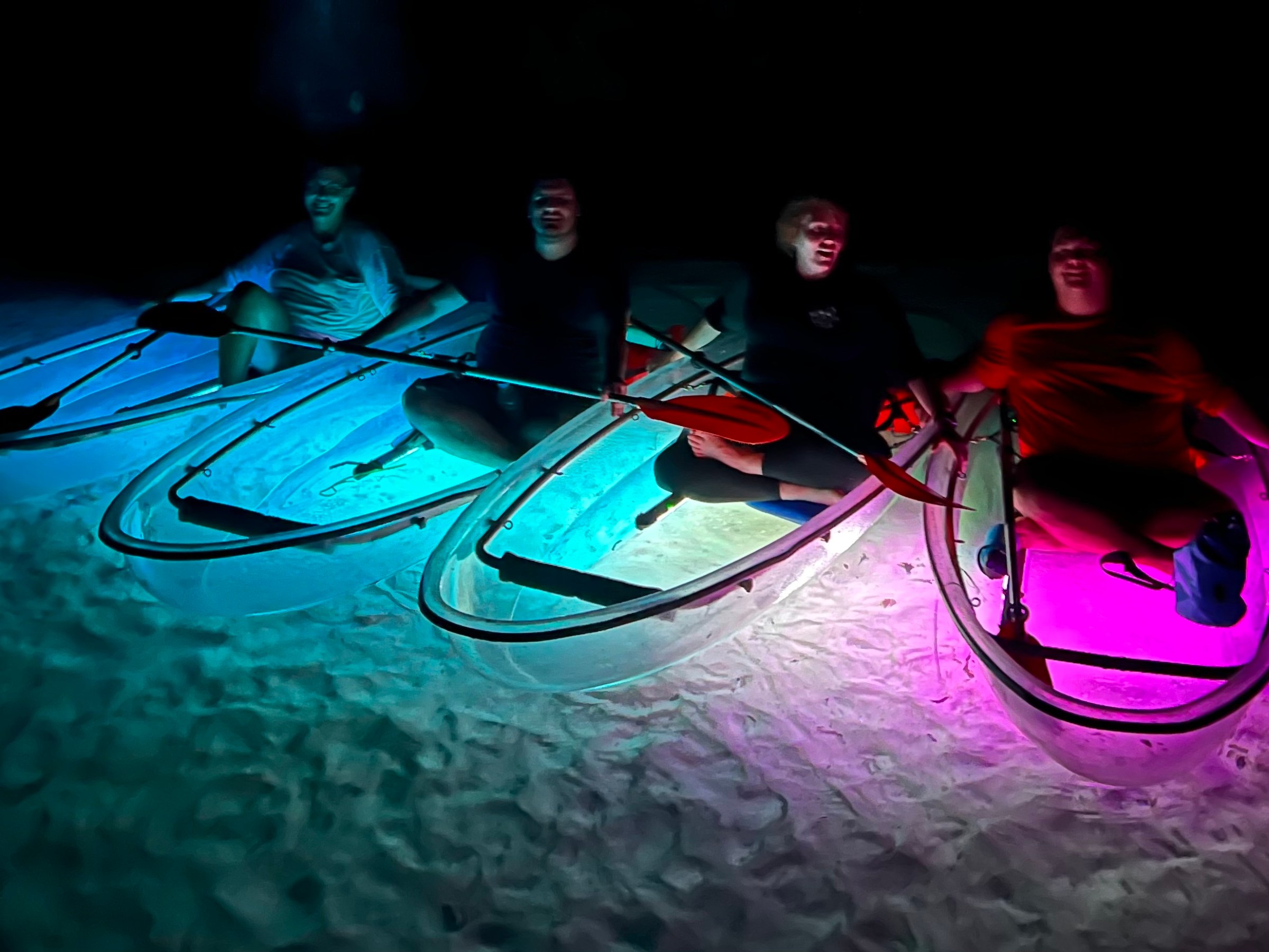 Four people sitting in illuminated clear kayaks on sand at night.