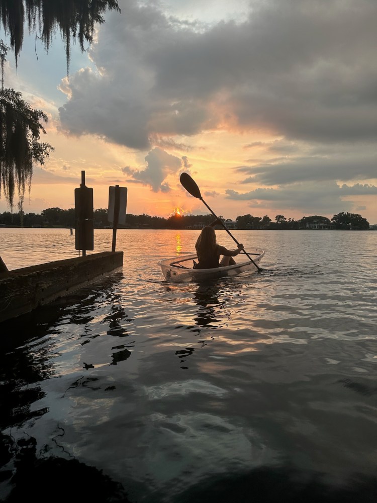 Person kayaking on a lake at sunset with clouds and silhouetted trees.