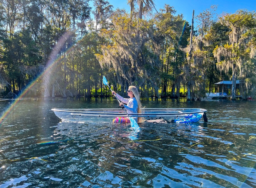 Person kayaking on clear water with trees in the background and a rainbow reflection on the water.