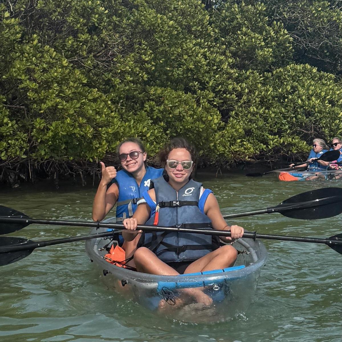 Two people kayaking in clear kayak, near mangroves, wearing life vests and sunglasses, one giving thumbs up.