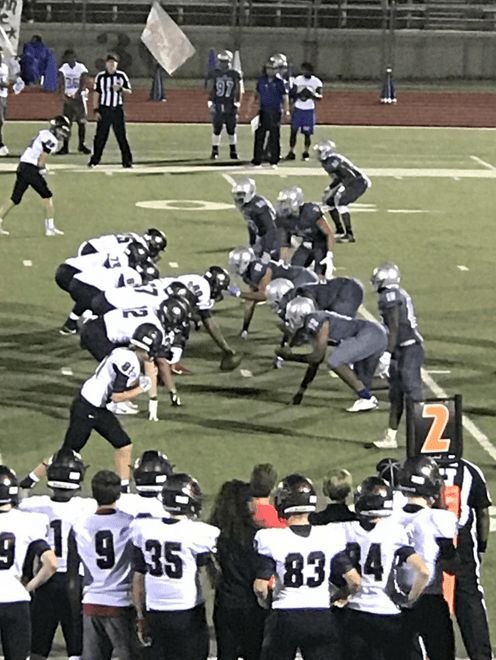 Two football teams in formation on a field ready for a play, with players on the sideline observing.