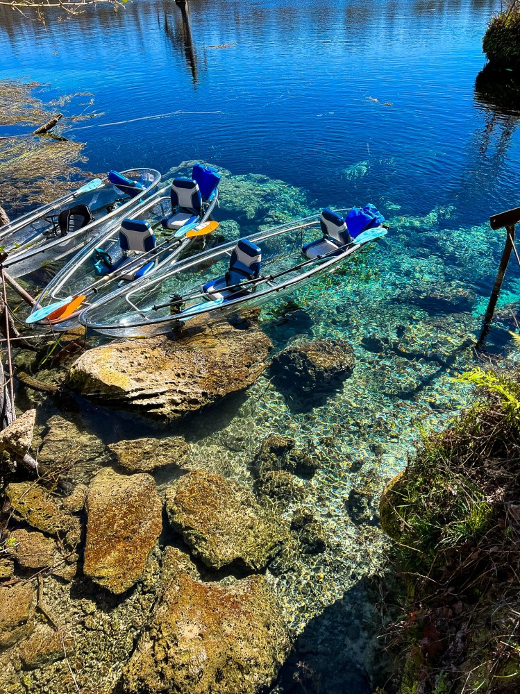 Three clear kayaks floating on a crystal-clear blue water lake with visible rocks.