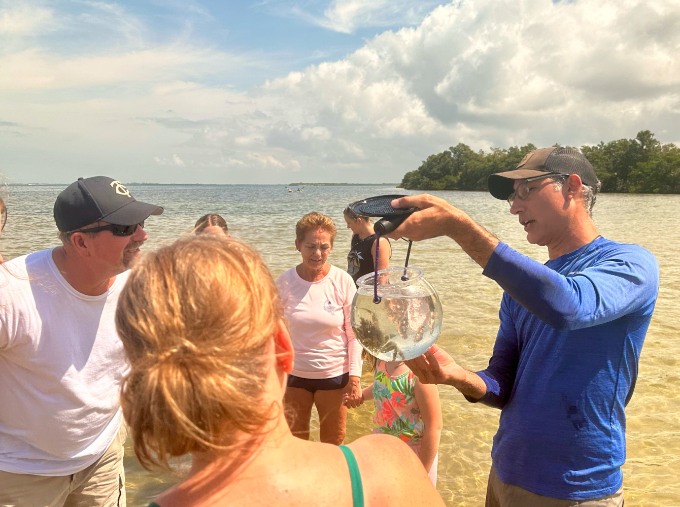 Group gathered on beach, man holding a glass container, discussing contents, sea in background.