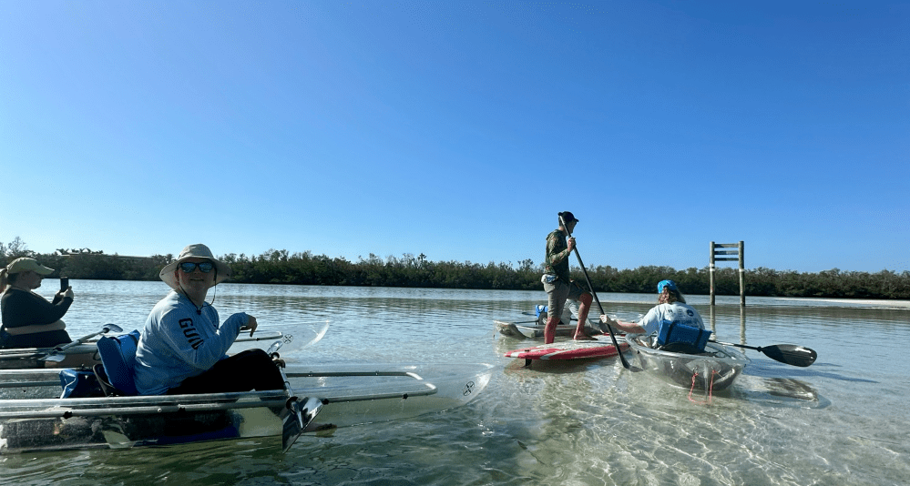 People kayaking and paddleboarding on clear shallow water under a blue sky.