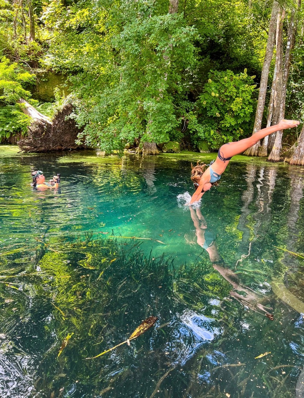 Woman diving into a clear lake surrounded by trees, with another person in the water.