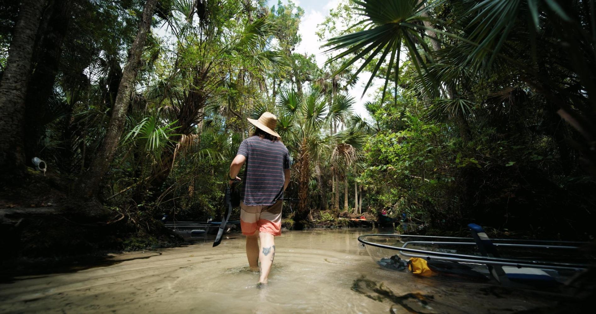 Person in hat walking through shallow water in a forest with kayaks nearby.