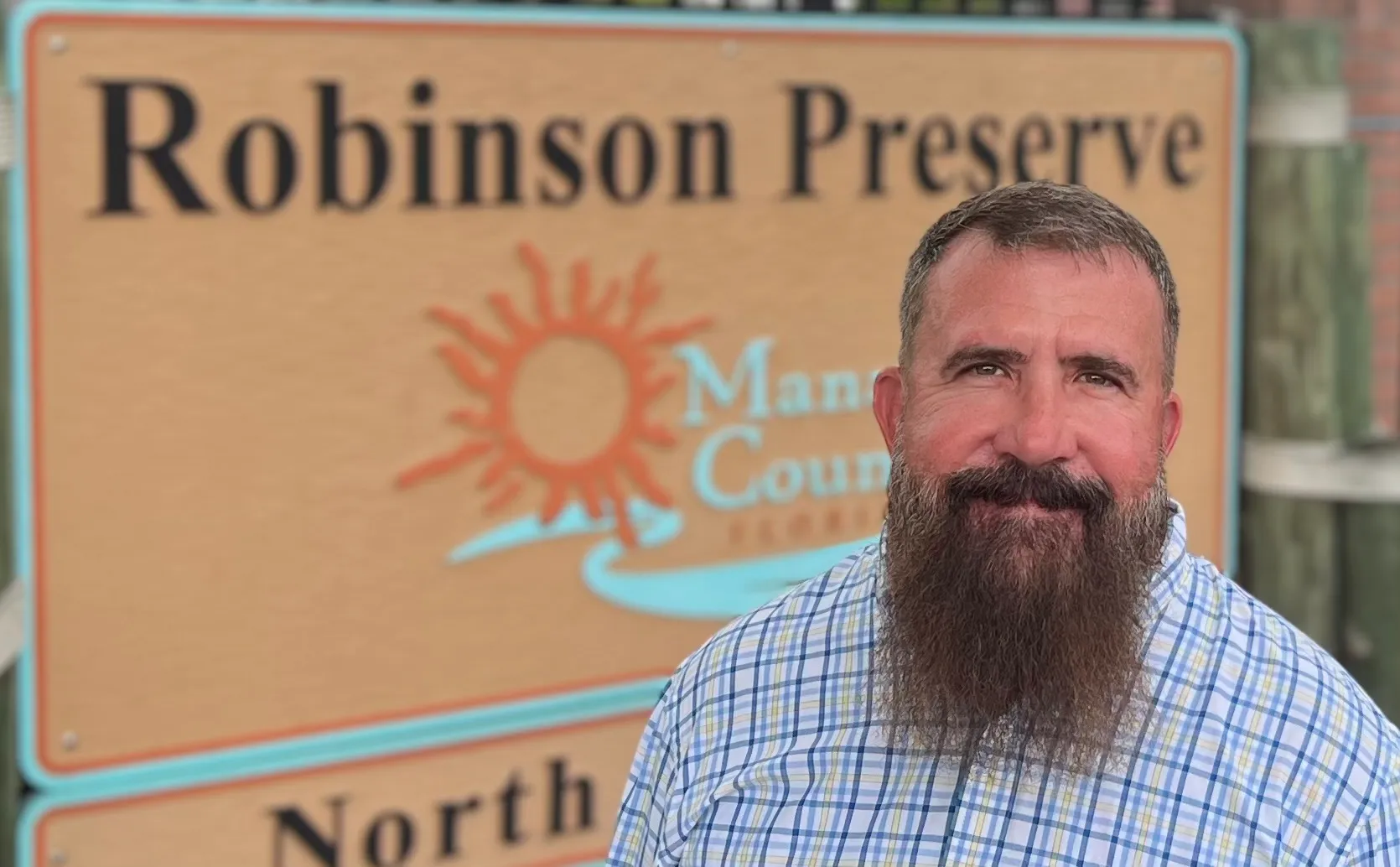 Person with a beard in a checkered shirt stands in front of a Robinson Preserve sign.
