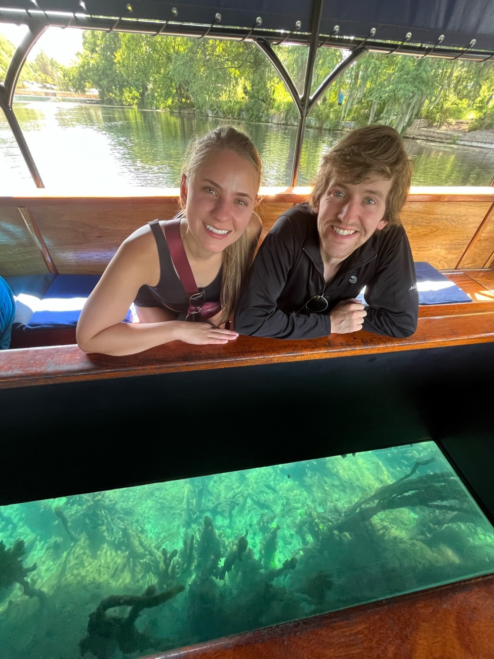 Two people smiling on a boat with a clear glass bottom displaying underwater view.
