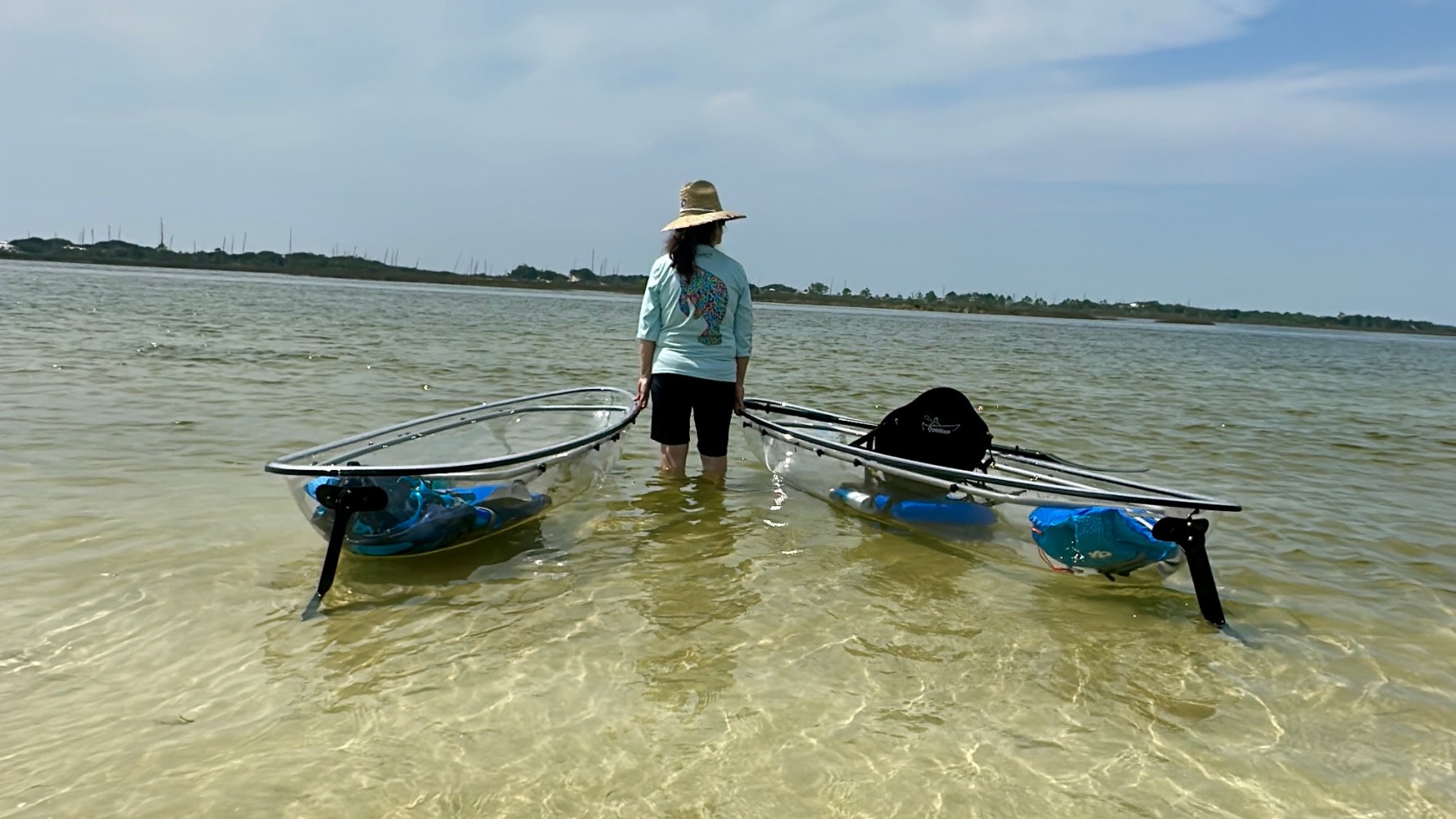 Person with hat stands in water holding two transparent kayaks.