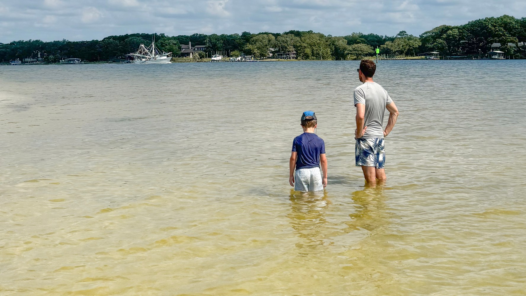 Two people standing in shallow water facing a lake with boats and trees in the background.