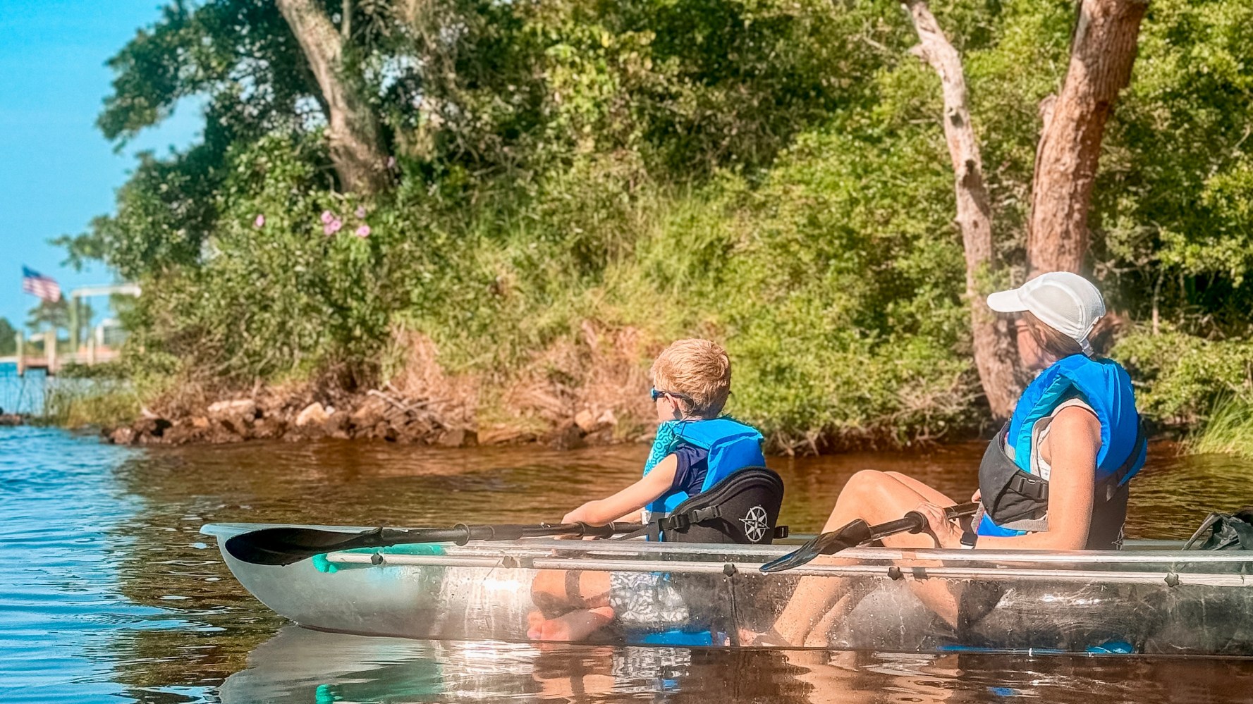 Two people in life jackets kayaking on a river with trees and an American flag in the background.