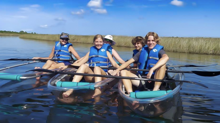 Four people in blue life vests sitting in clear kayaks on calm water.