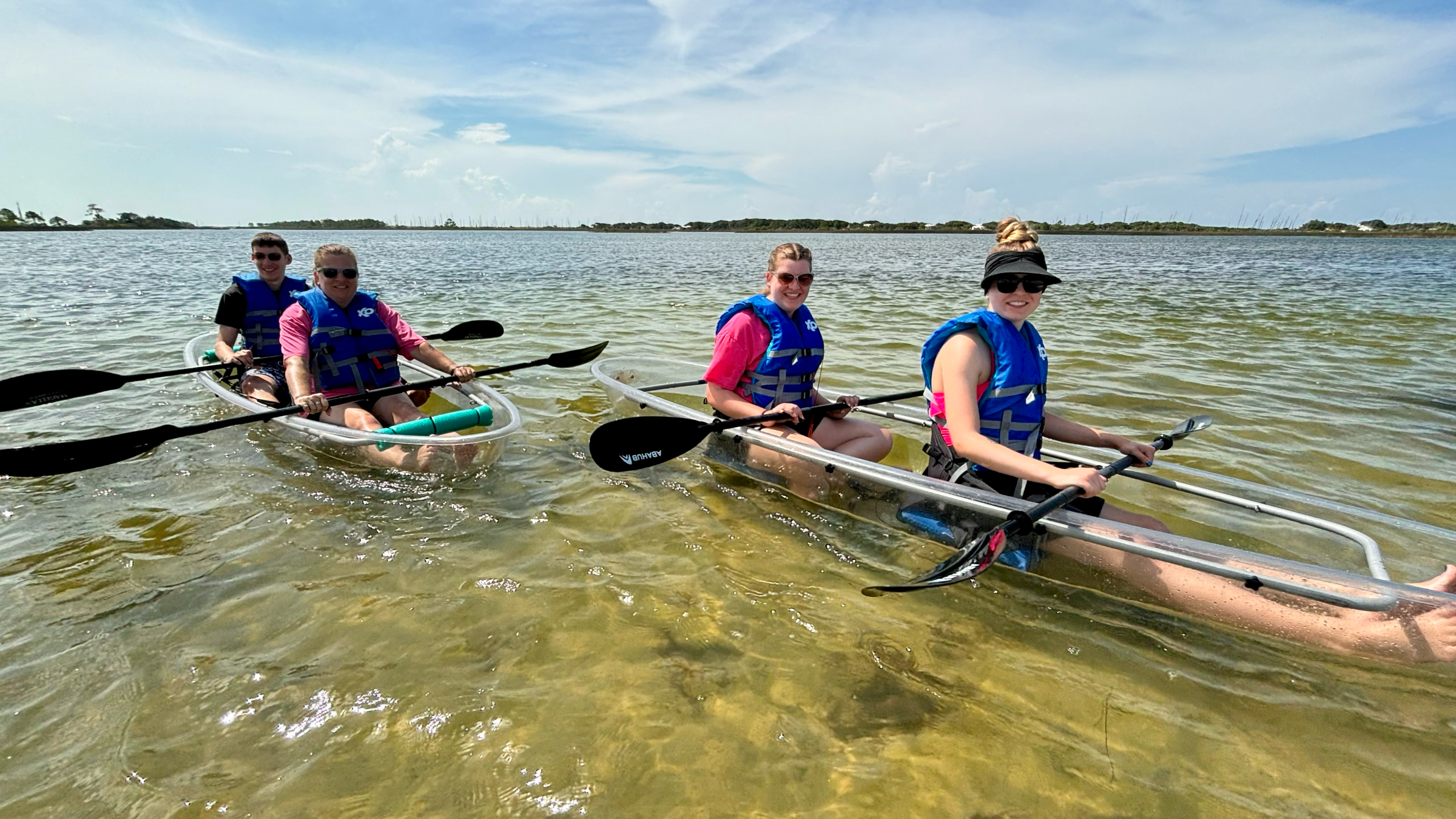 Four people in clear kayaks on a sunny day, wearing life vests and paddling on calm water.