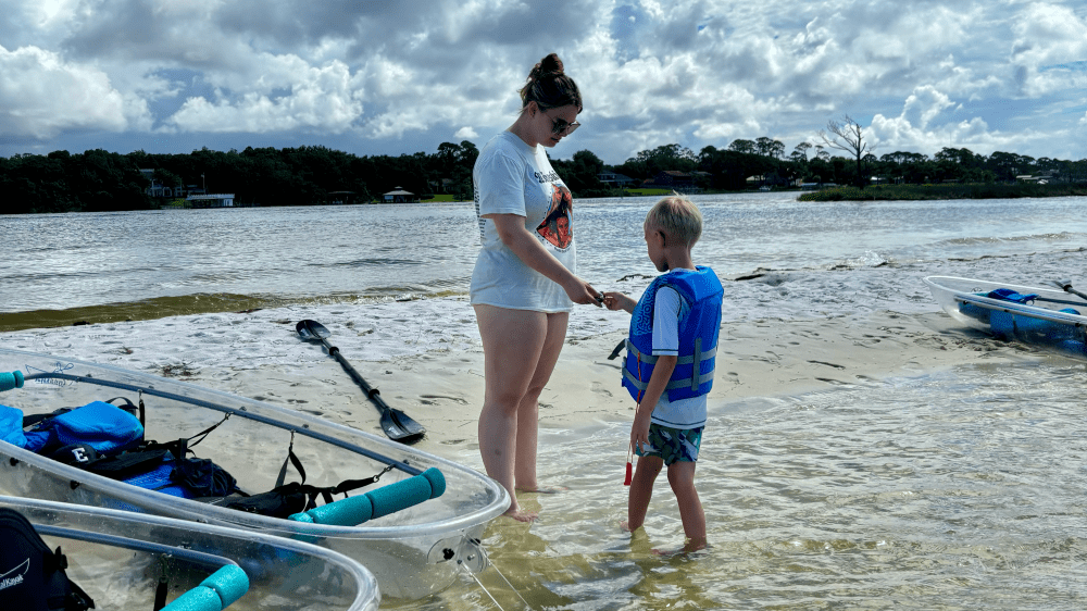 Woman and child with life vests by kayaks on a sandy riverbank.
