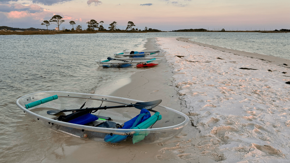 Transparent kayaks on sandy beach with trees and water at sunset.